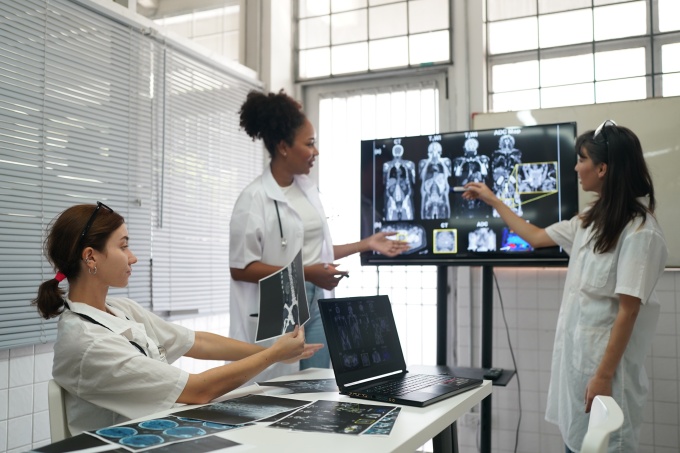 Three medical professionals discuss CT and MRI scans displayed on a large...